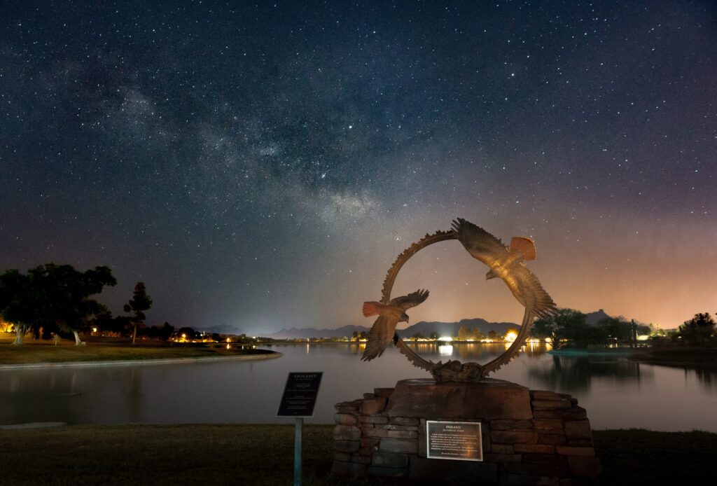 Image of night sky above bronze statue of circling raptors in front of a lake.