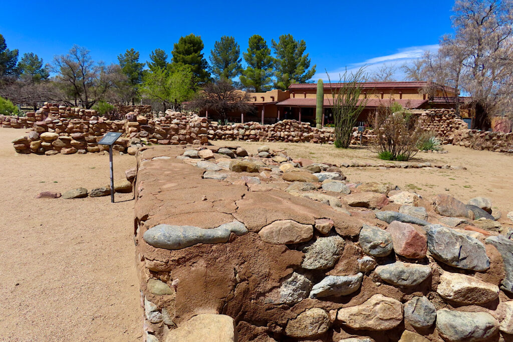 Rock walls covered in reddish brown plaster in front of low red-roofed building and line of dark green trees under blue sky.