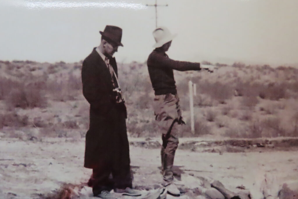 Black and white photo of man in dark hat and long dark coat standing beside a woman in tall leather boots, light-coloured pants, dark sweater and floppy hat pointing at something outside of image.