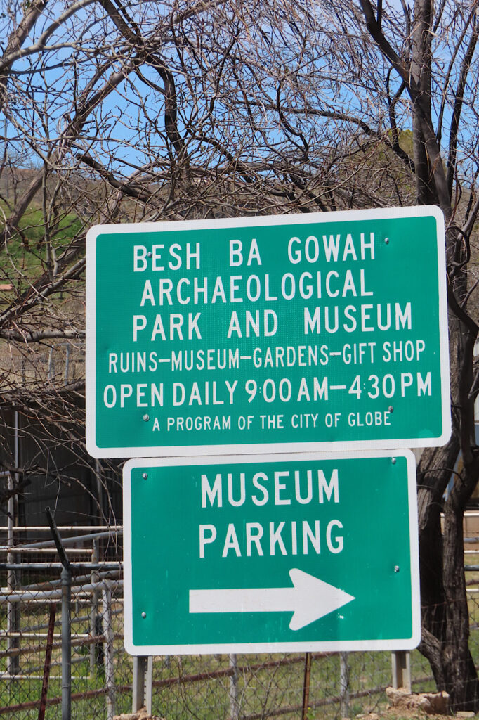 Two green sign swith white lettering. Top sign reads: Besh Ba Gowah Archaeological Park and Museum. Ruins-Museum-Gardens-Gift Shop. Open daily 900 AM - 430 PM. A program of the city of Globe. Bottom sign reads: Museum Parking (with a white arrow pointing to the right).