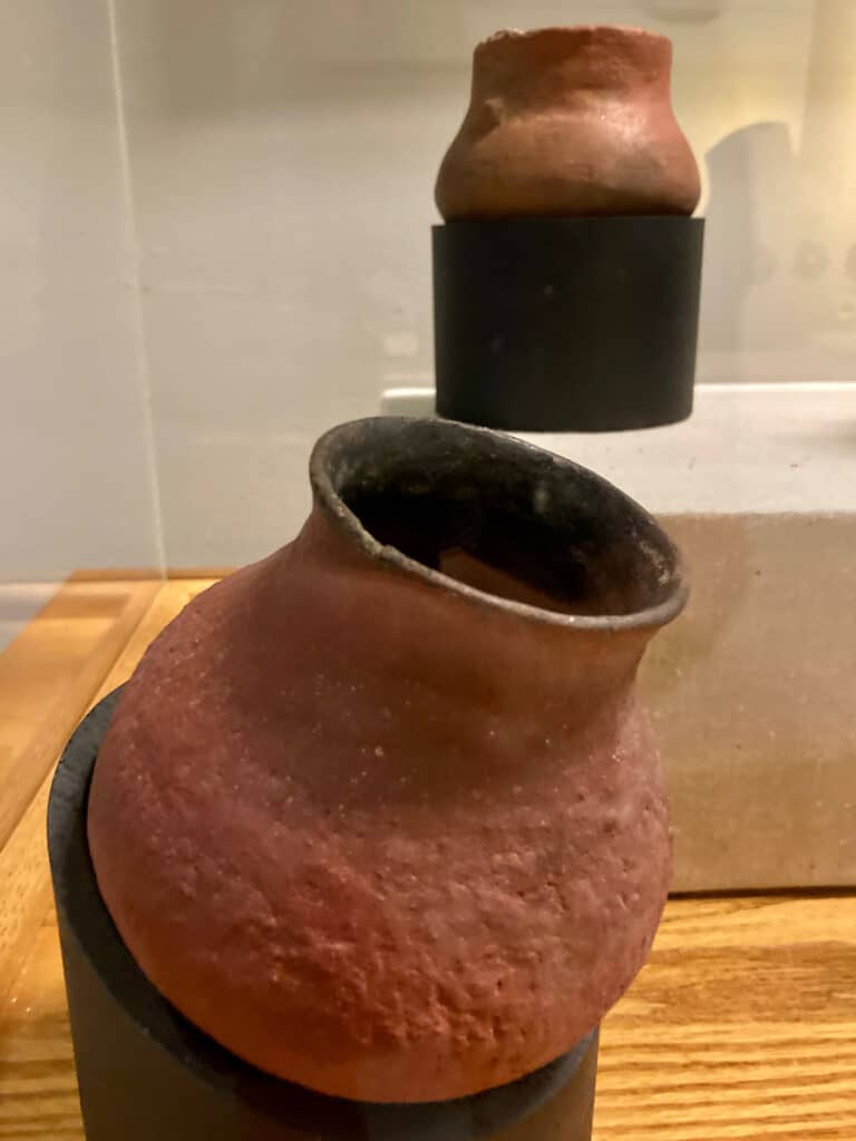 Large rustic red pottery jar in foreground of display case with smaller jar in background.