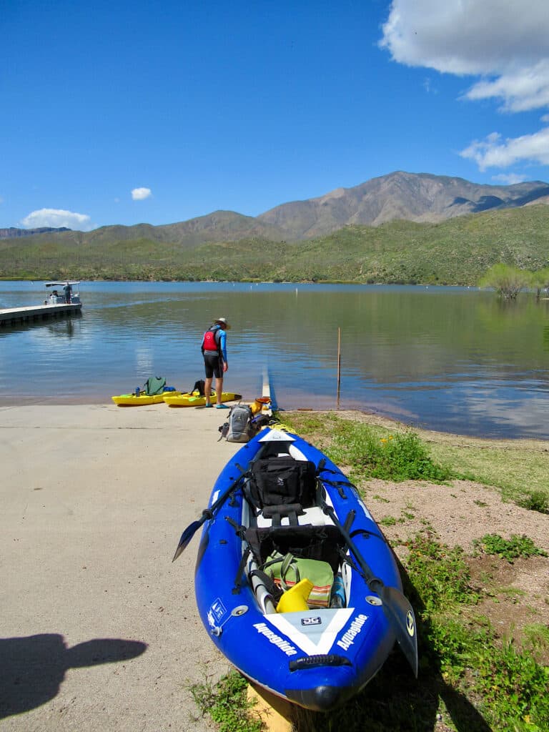 Man in shorts and red life jacket standing by two yellow sit-on kayaks by water's edge with inflatable blue and white kayak sitting in foreground.