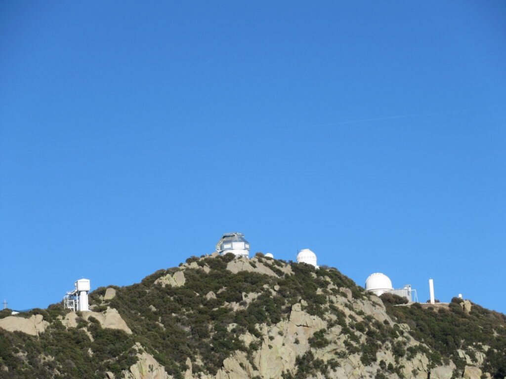 Five white observatories of varying shapes on top of a rocky, partially-brush covered peak under blue sky.