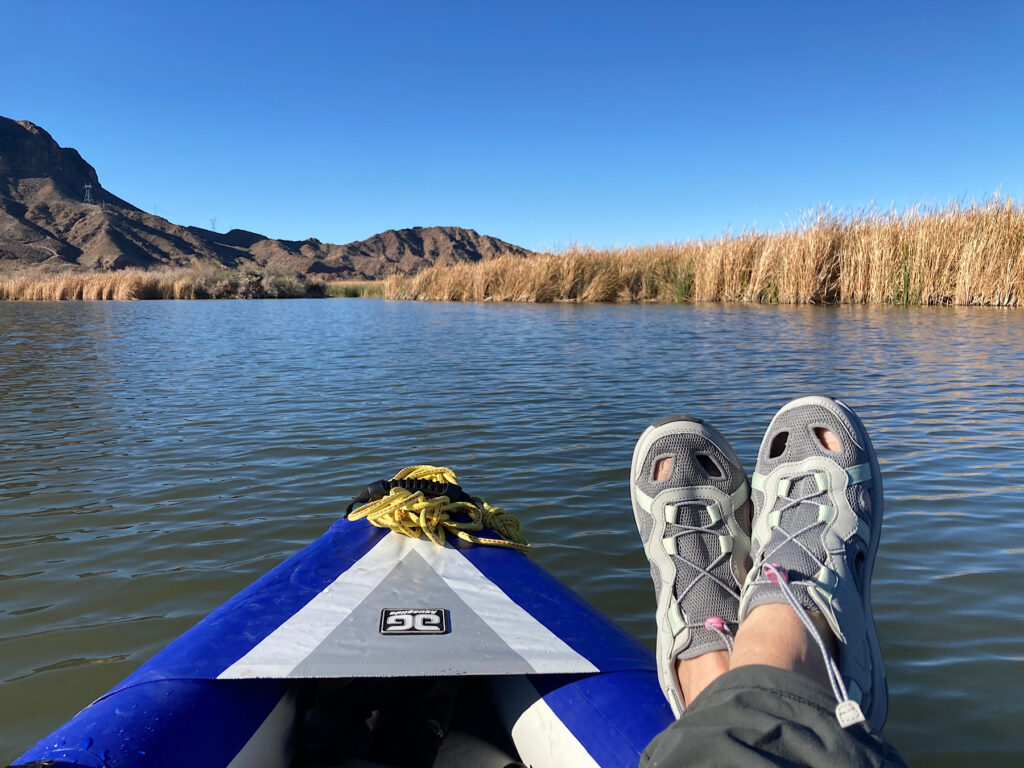 Woman's feet crossed on bow of inflatable blue and white kayak on waterway lined with reeds under blue sky.