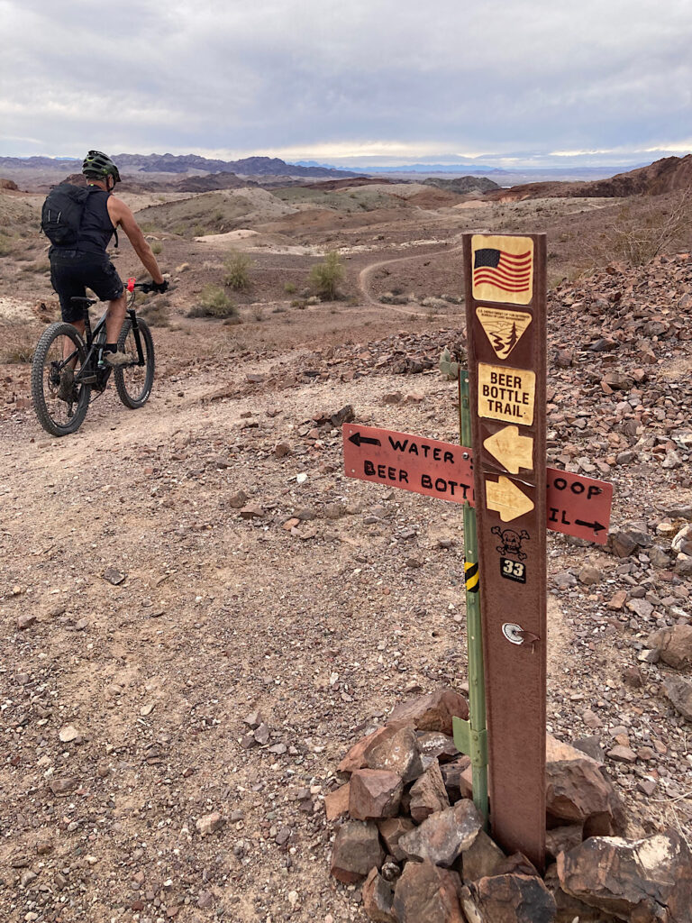 Man on mountain bike behind cross-shaped brown trail sign reading "Beer Bottle" on top of "Watershed."