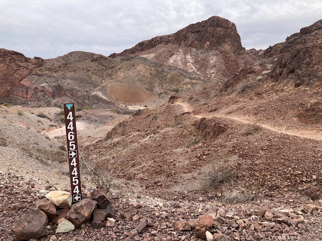 Brown trail markers with white numbers marking trail junction below ragged brown cliffs.