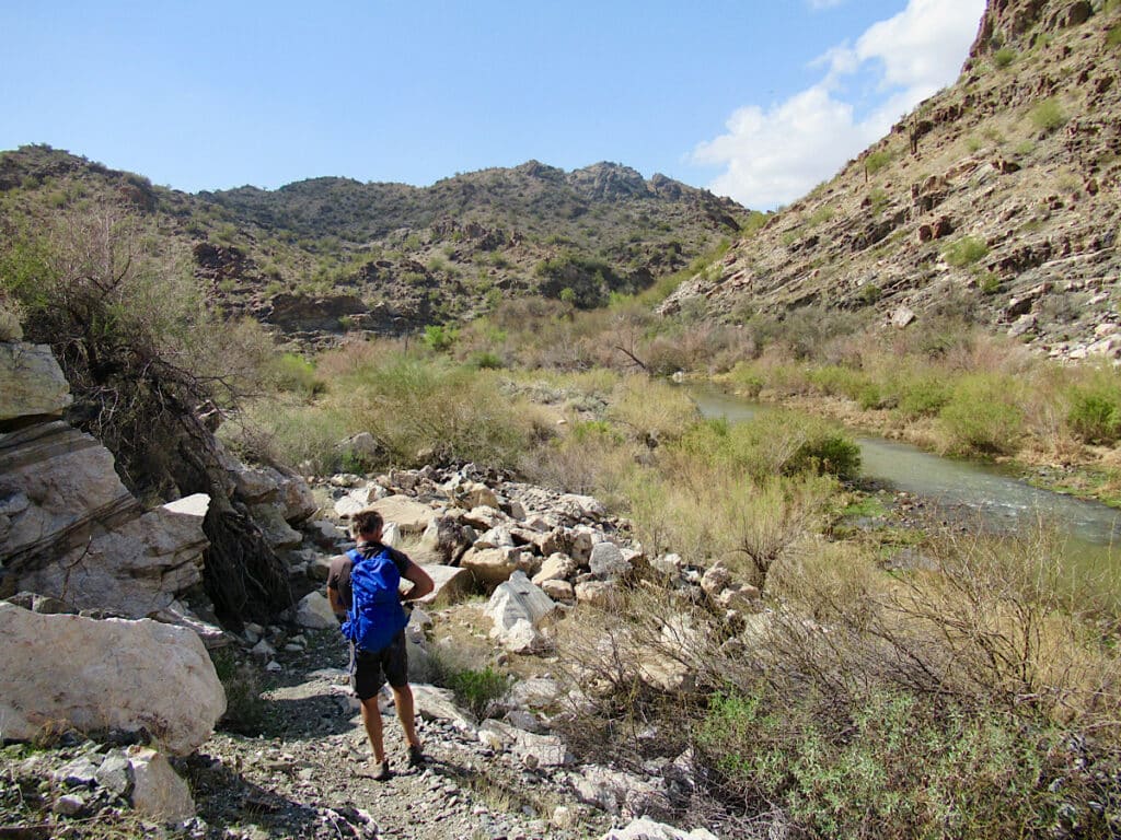 Man in shorts carrying blue backpack walking old roadway above green river valley.