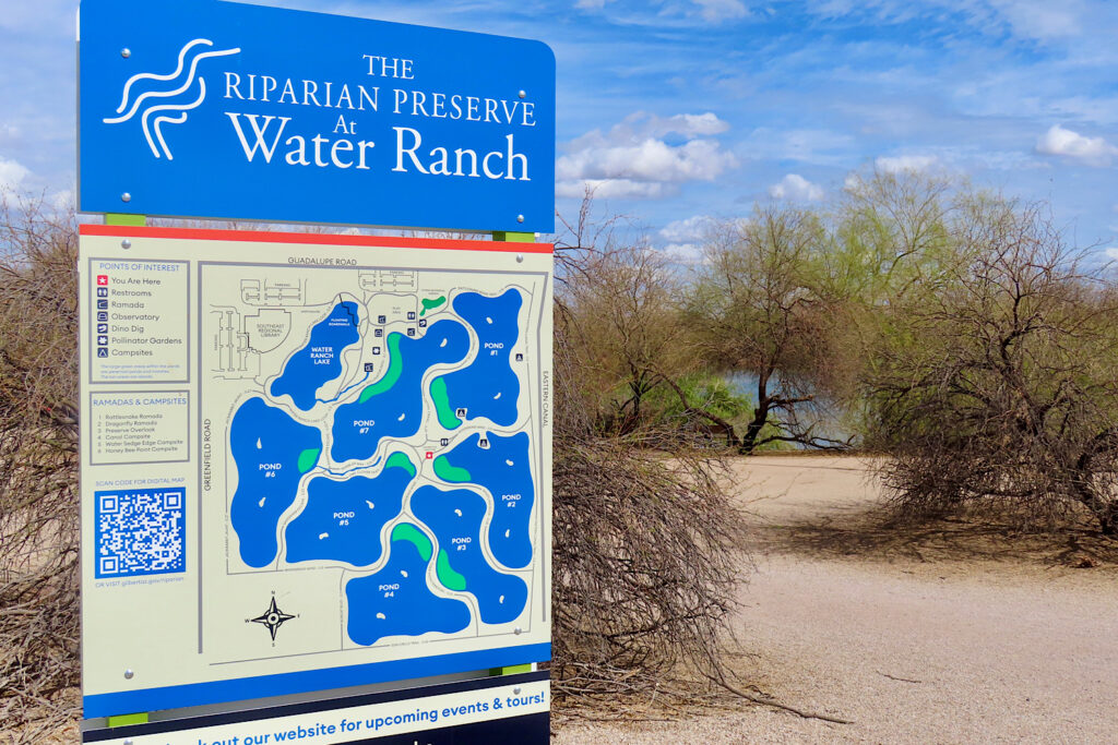 Blue, pale yellow, and green map sign of Gilbert Water Ranch in front of dirt trail leading to water.