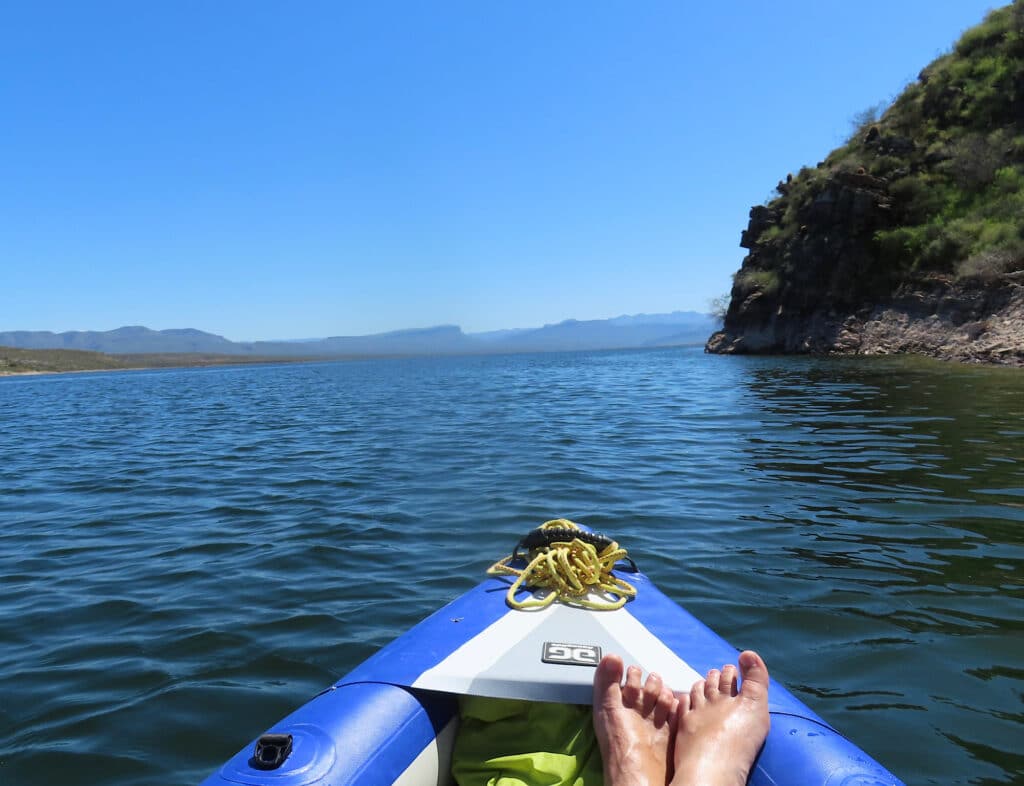 Woman's bare feet propped up on front of blue and white inflatable kayak on lake near island.