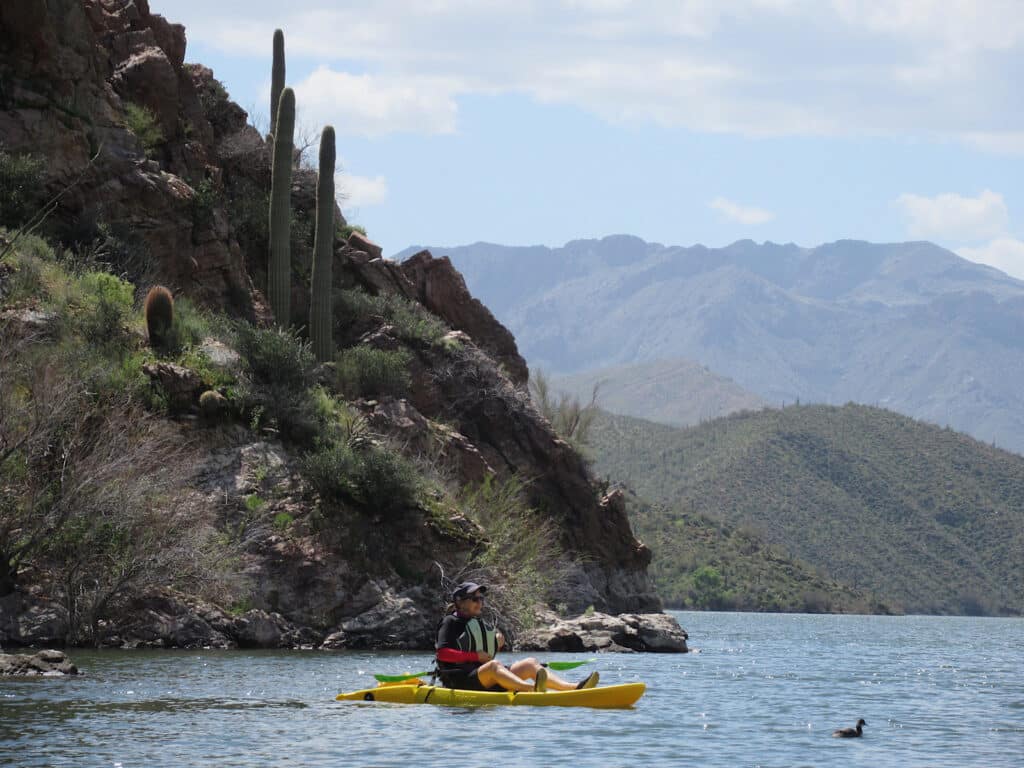 Woman on yellow sit on top kayak in lake with small bird in front beside steep cliff.