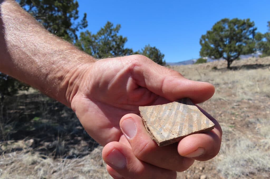 Man's hand holding beige pottery sherd with black painted stripes.