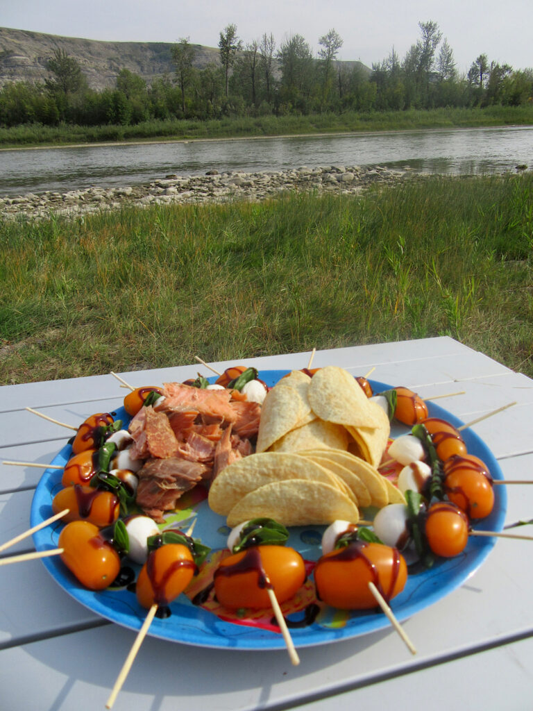 Tomato cheese skewers surrounding chips and smoked salmon sitting on a plate on a metal table by a river.