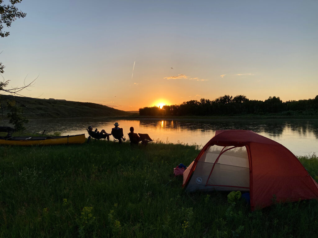 Silhouette of people sitting in chairs by river with canoe and tent in foreground. Distant sunset.