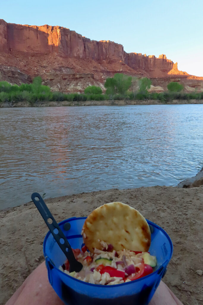 Blue bowl with rice dish and a piece of naan balanced on legs of person sitting by river.