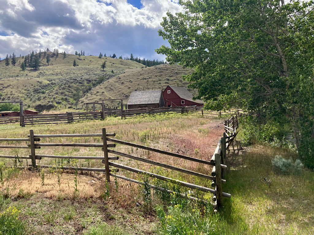 Wooden fence and field in front of distant barns and buildings at base of low hill.