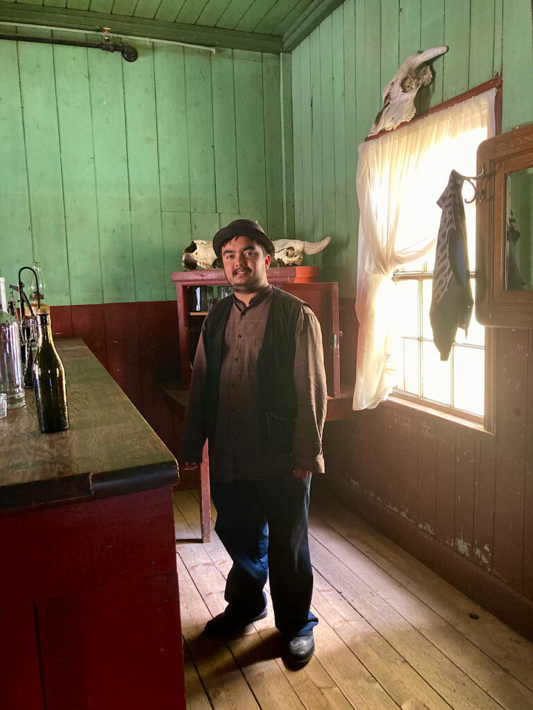 Man in historic clothing standing behind a bar with green painted walls and a window behind him.