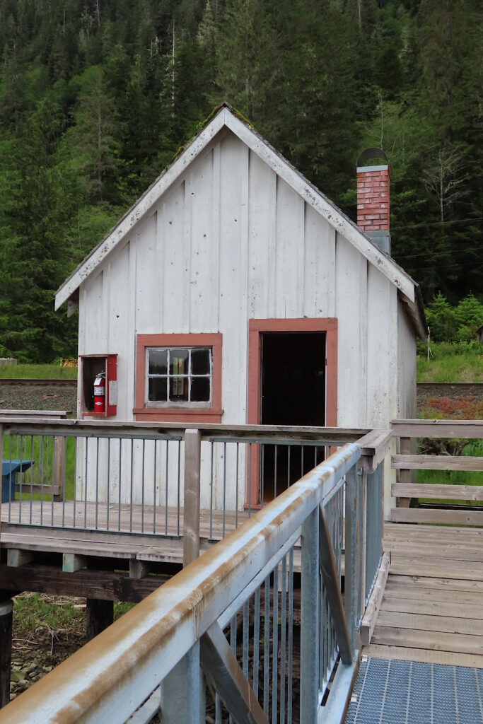 Small white building wiht a chimney, door and window on a wooden walkway.