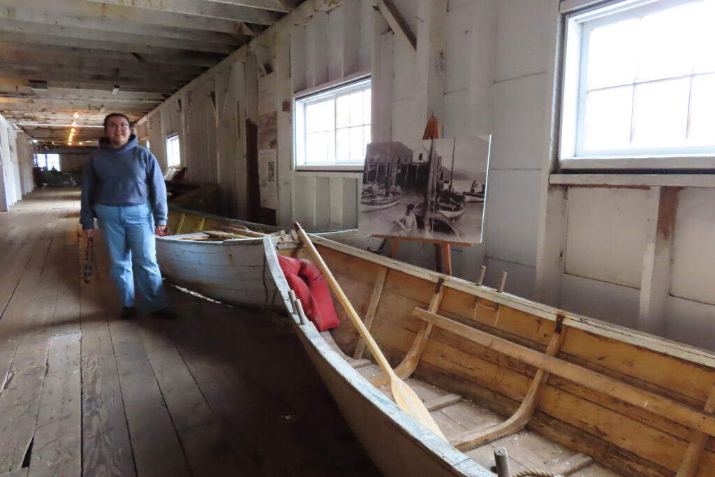 Woman in jeans and long sleeved shirt standing inside a buildling beside a wooden boat.