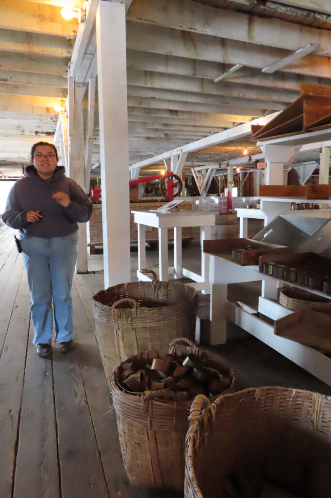 Woman in jeans and long sleeved shirt standing beside baskets holding metal tins.