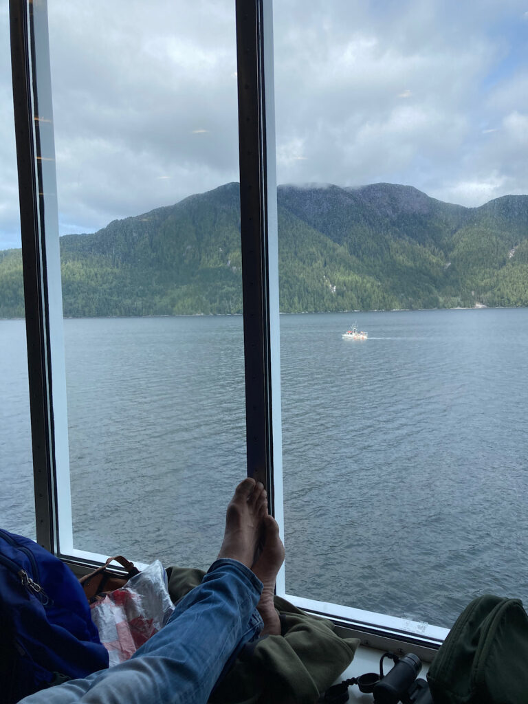 Man's feet resting on pack in front of picture window overlooking water and distant land.