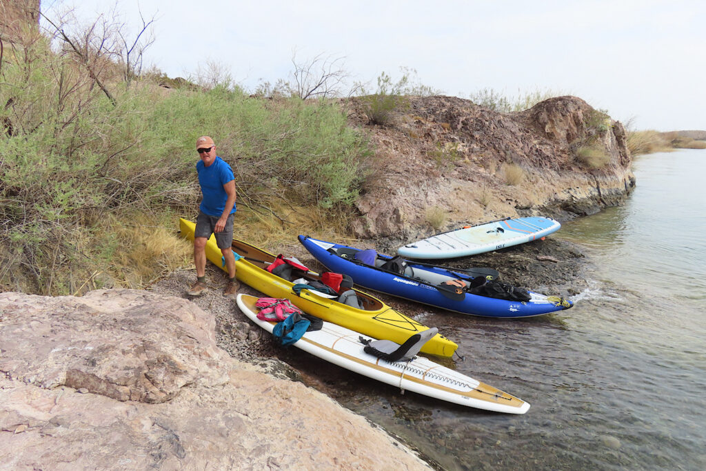 Man walking on shore in front of inflatable kayak and paddleboards sitting at water's edge.
