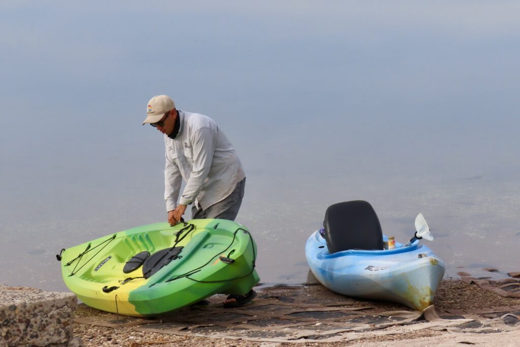 Man in hat and long sleeve shirt and pants moving green and yellow kayak. Another blue kayak nearby on shore.