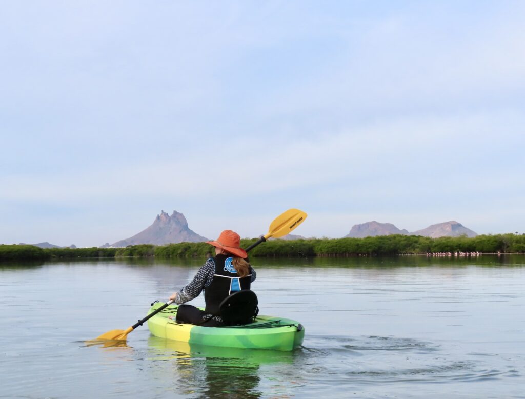 Woman wearing orange hat in green kayak on the water with distant mountains.