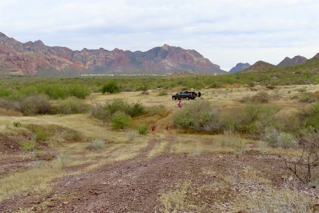 Truck parked in grassy meadow with hikers heading up red rock road and mountains in distance.