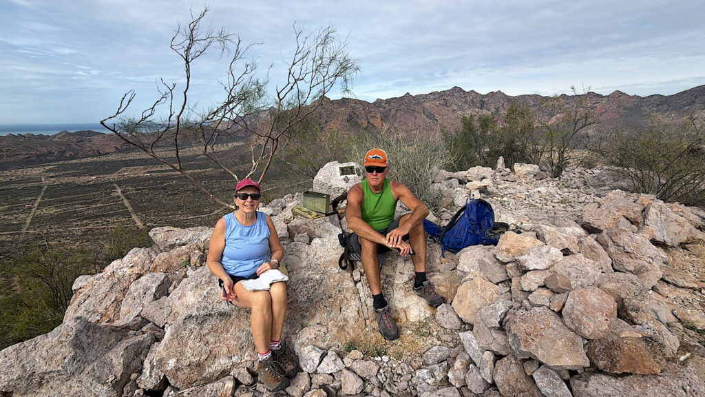 A man and woman on a rocky peak with a notebook in the woman's lap and memorial plaque behind.