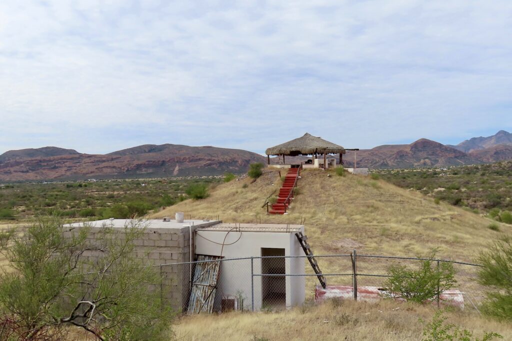 Thatched building on small hill surrounded by chain link fence and outbuildings.