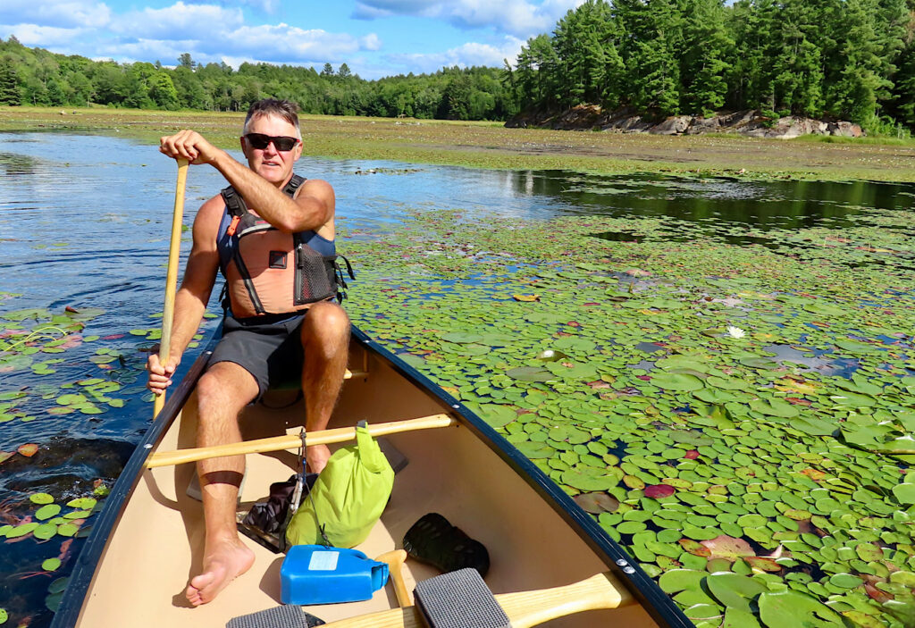 Man in back of canoe paddling through thick lily pads.