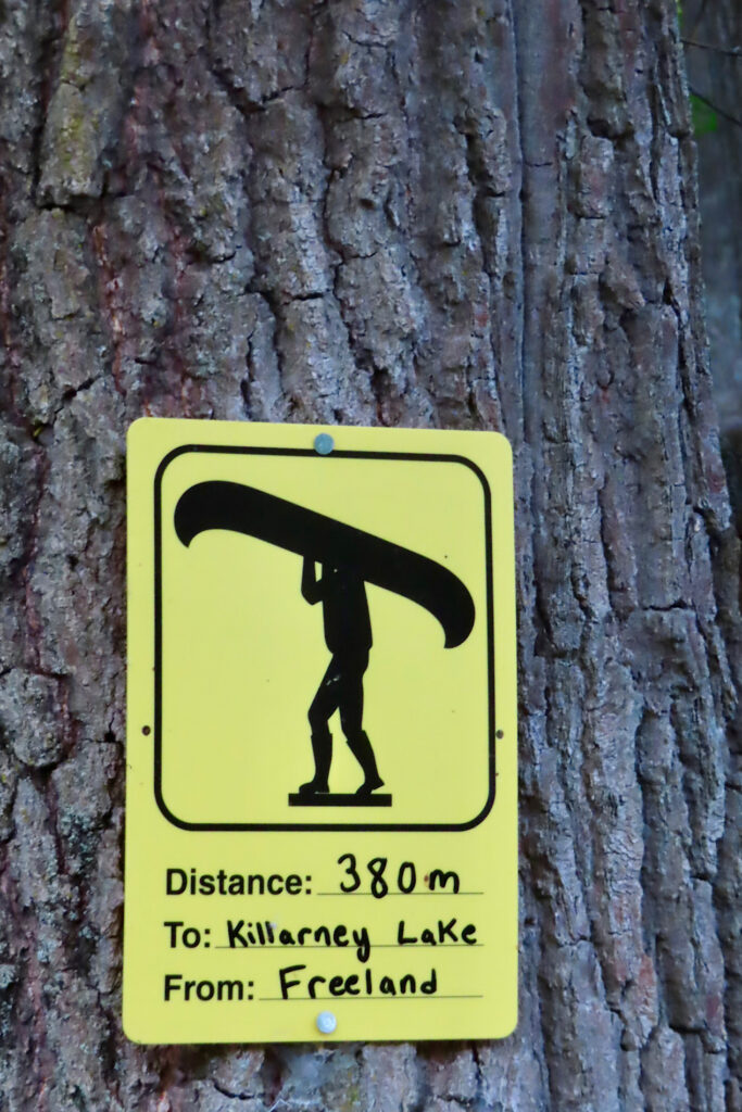Yellow sign with black image of person carrying a canoe overhead. Text reads: Distance: 380 m To: Killarney Lake From: Freeland