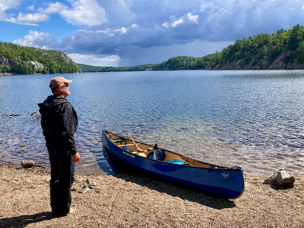 Man in black raingear and bare feet standing beside blue canoe pulled up on pebble beach.