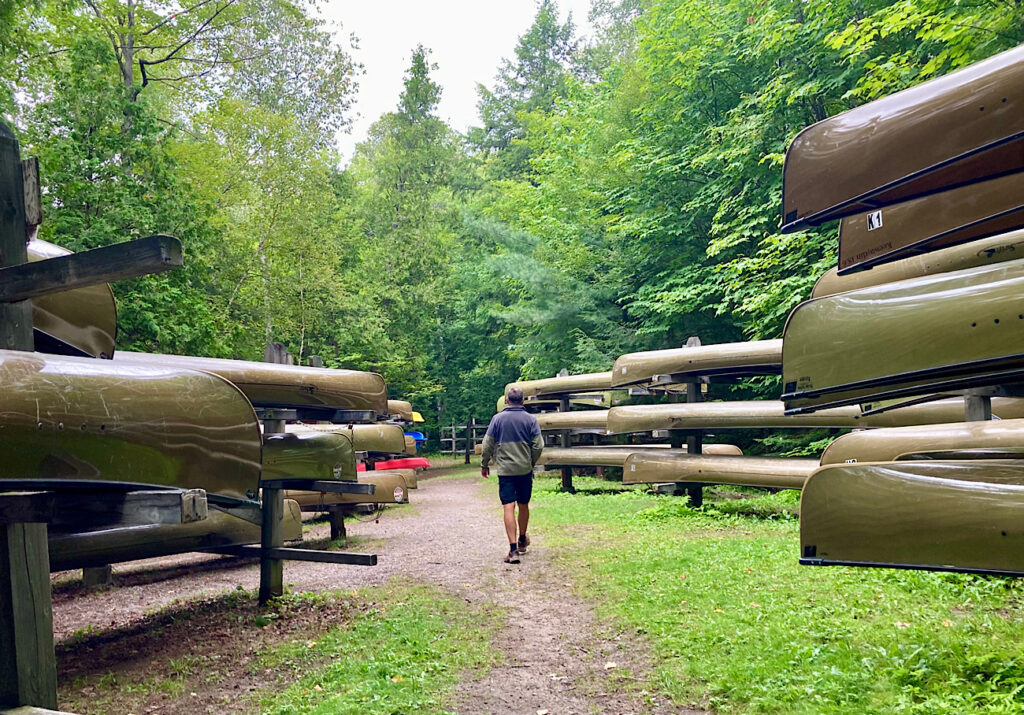 Man walking down path in deciduous forest with racks of light brown canoes on either side.