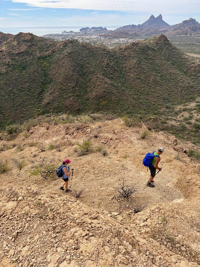Man and woman hiking down rocky slope above distant mountain and ocean.