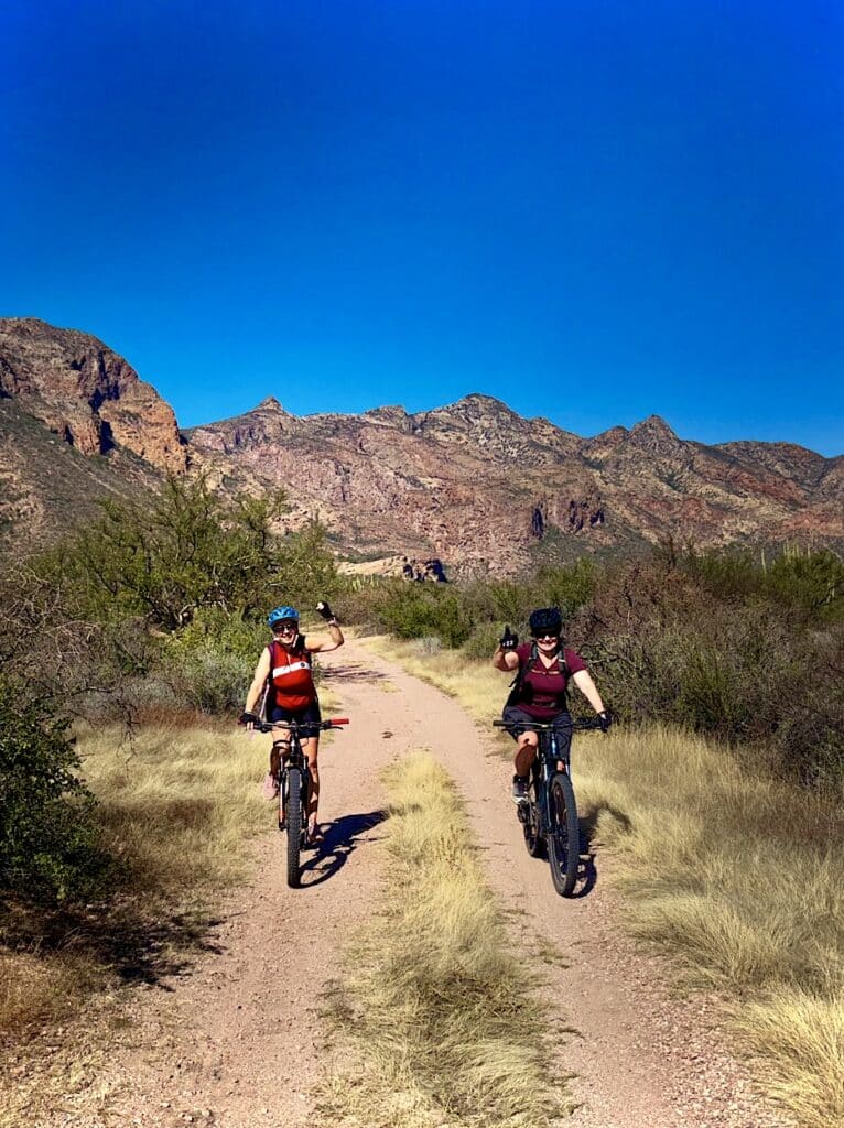 Two women on mountain bikes riding side by side with thumbs up on old road in desert.