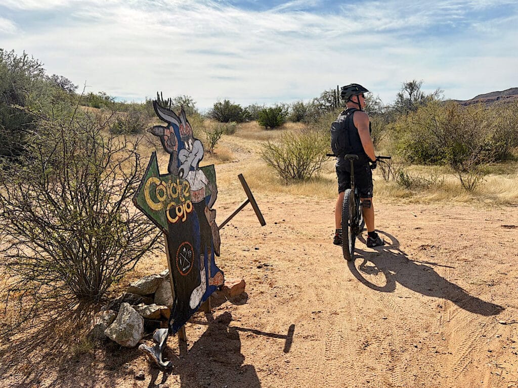 Man on mountain bike looking at phone next to cut out trail sign of coyote reading: Coyote Cojo.