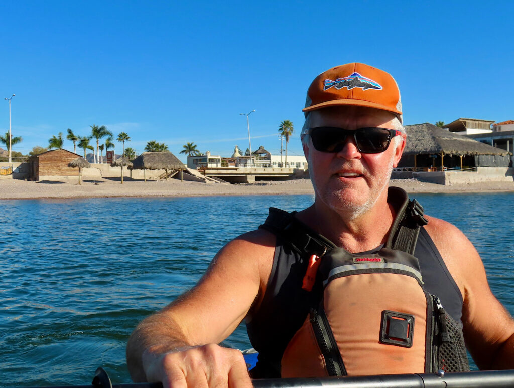 Man in orange life jacket and orange ball cap paddling in front of a beach lined with buildings.