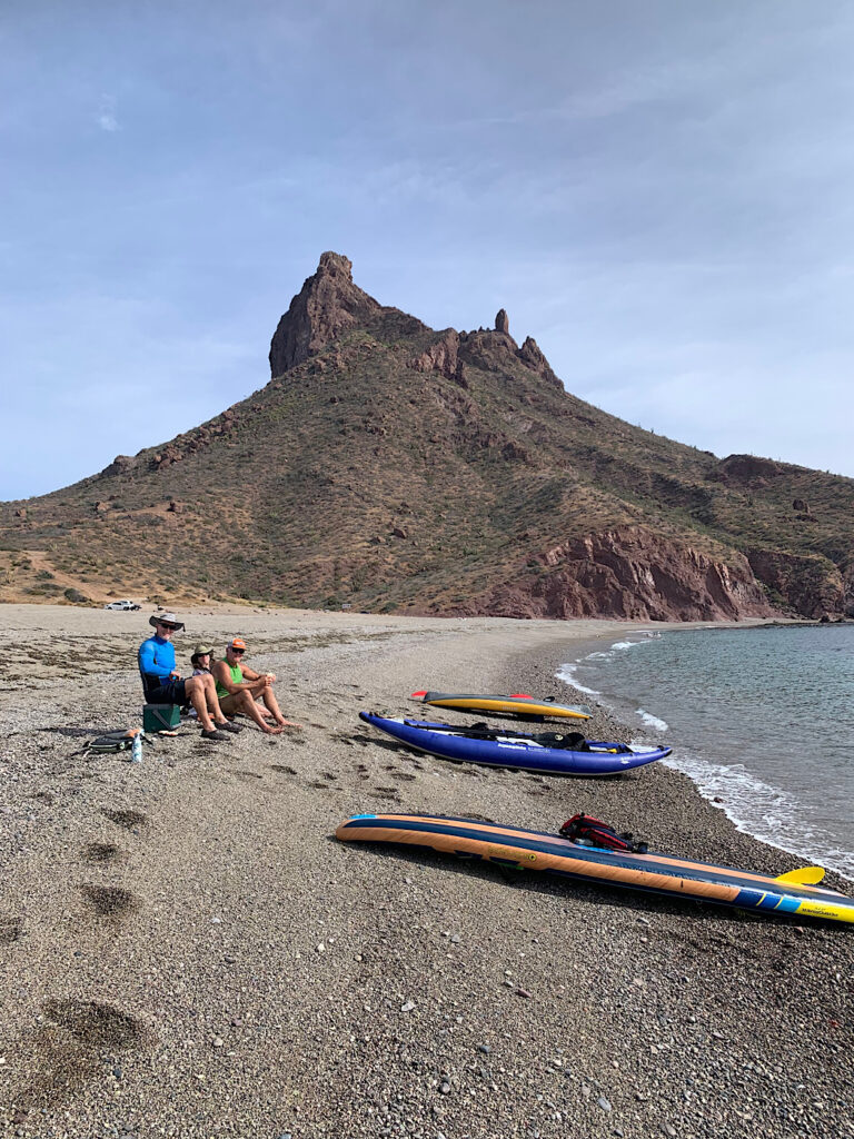 Three people sitting on pebble beach above kayaks.