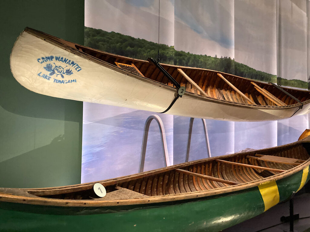 Two canoes on display, one with "Camp Wanapitei" stamped in blue on bow; the other painted green with two broad yellow stripes near center of boat.