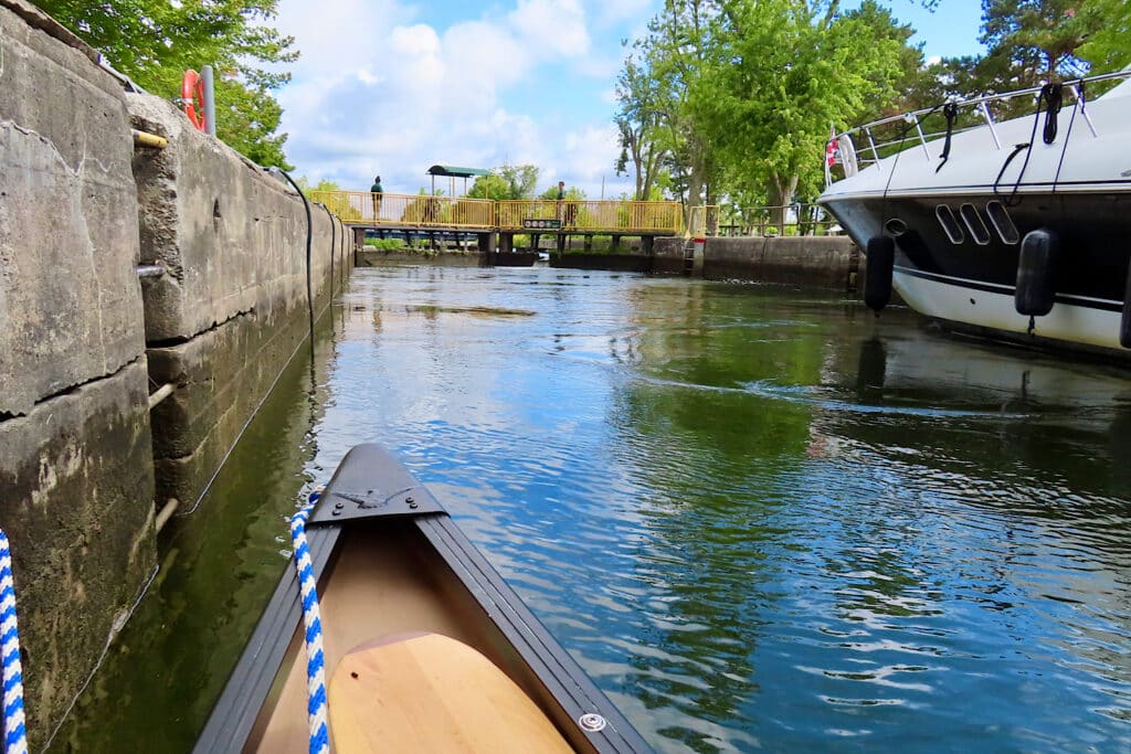 Bow of canoe in a pool of water beside a concrete wall with a cruising boat on opposite side of passage.