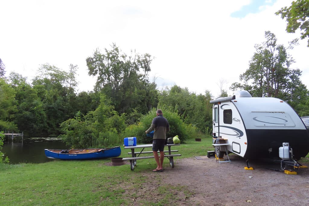 Man at picnic table between a small white trailer and a blue canoe perched at water's edge.
