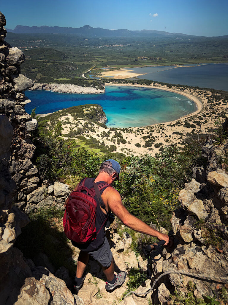 Man wearing red backpack hiking down break in rock wall above blue beach.