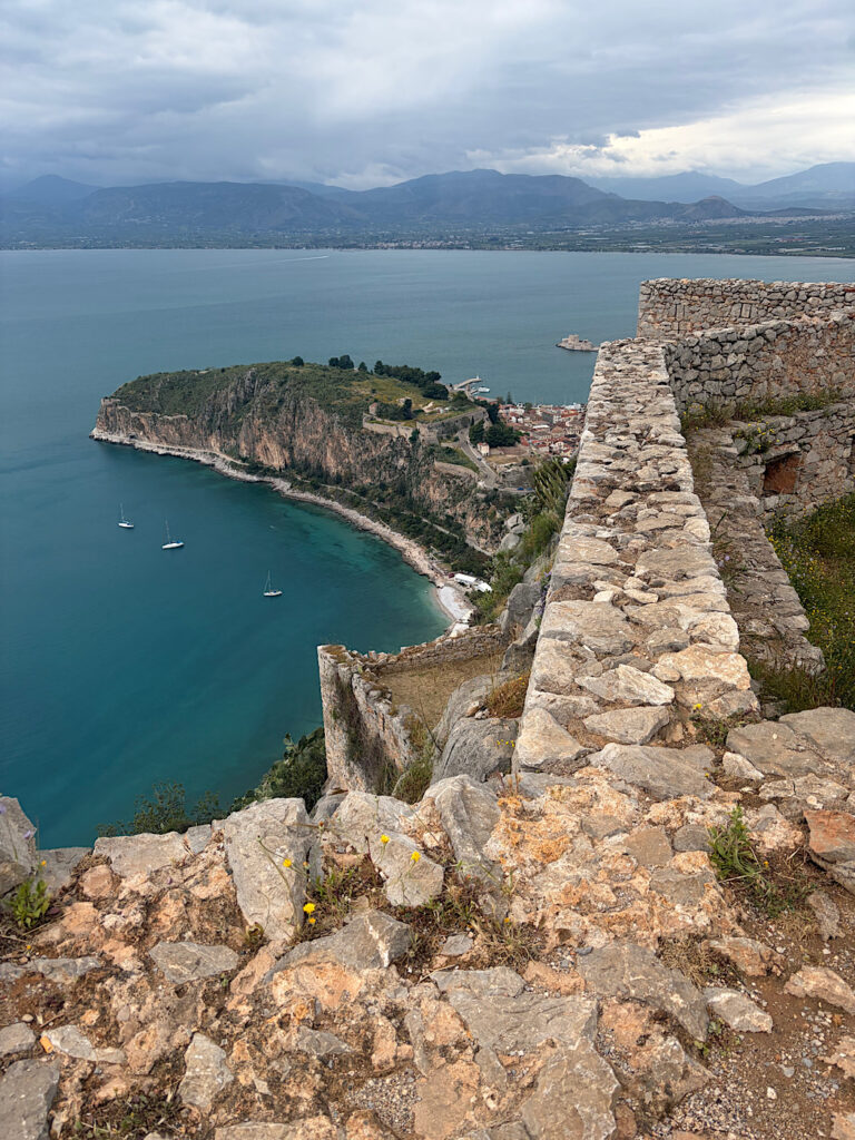 View of stone wall high above lower ruins and a town with a small island castle in distance.
