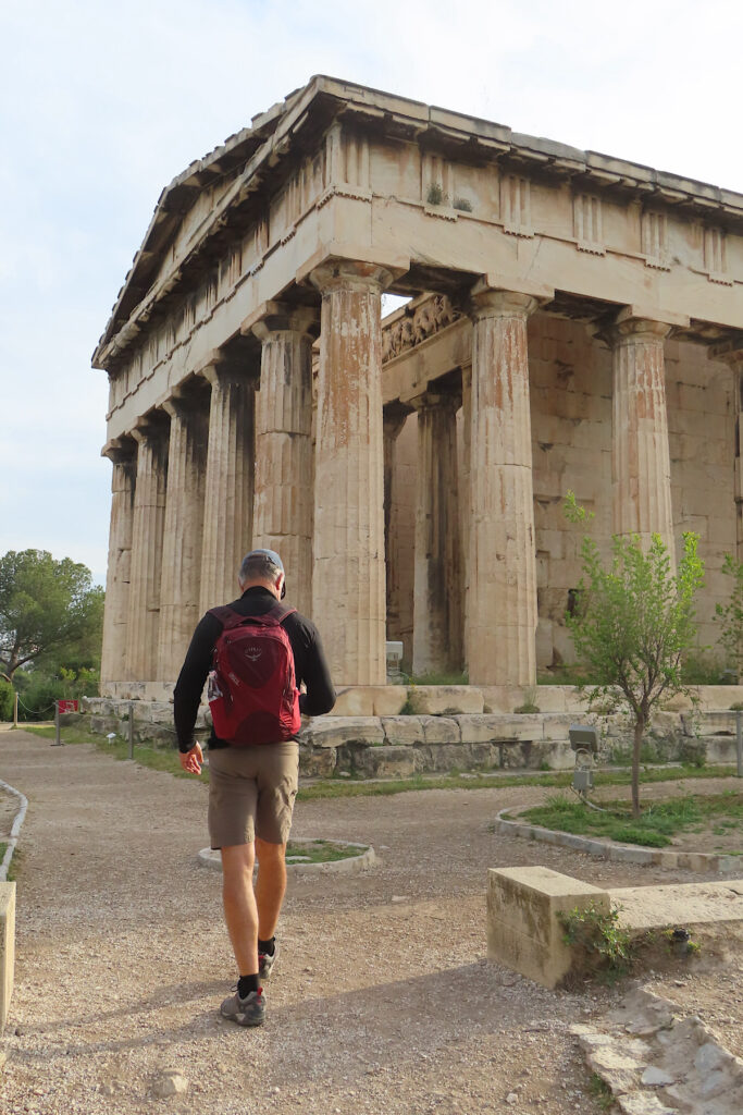 Man with red pack in shorts and long sleeve shirt walking toward large building with many columns.