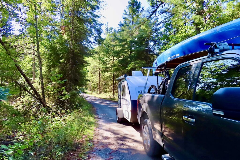 Truck with canoe and small trailer driving gravel road through forest.