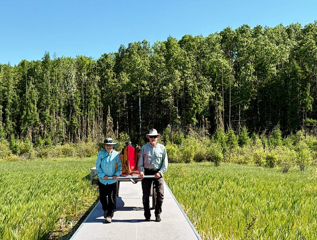 Woman and man hauling canoe cart on metal boardwalk in swampy area.