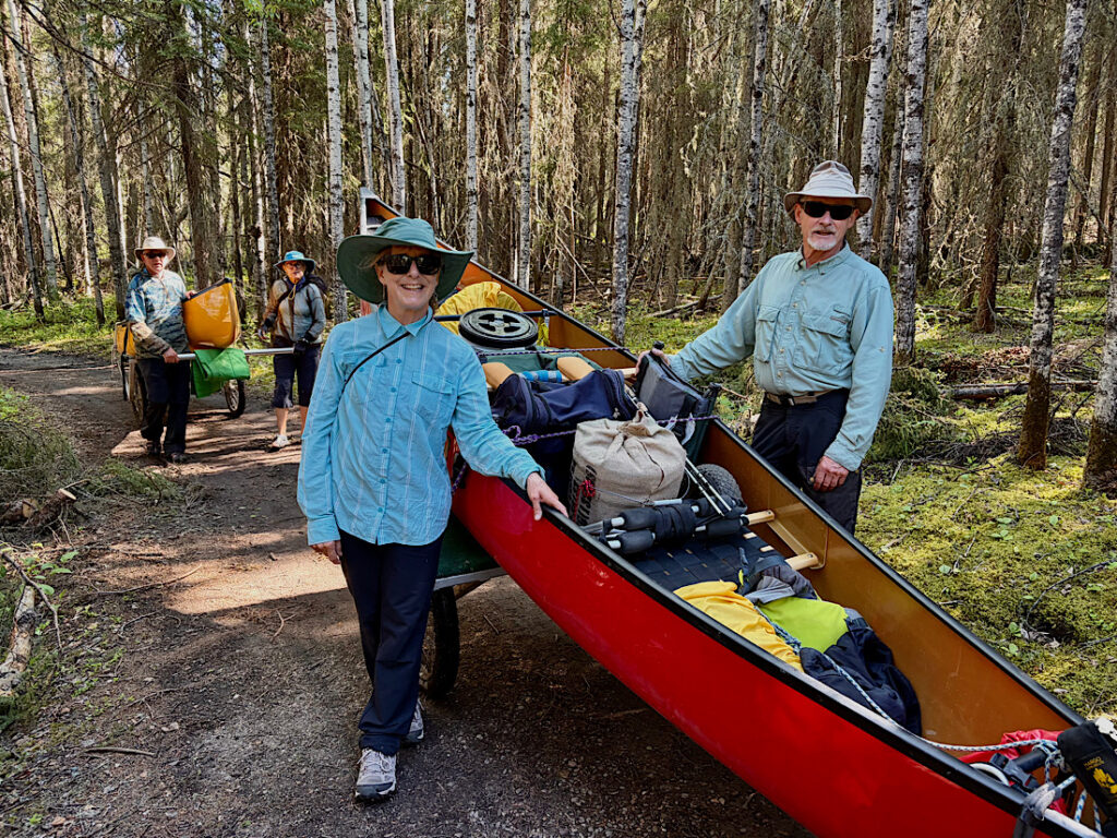 Man and woman standing beside loaded red canoe with another couple behind with yellow canoe.