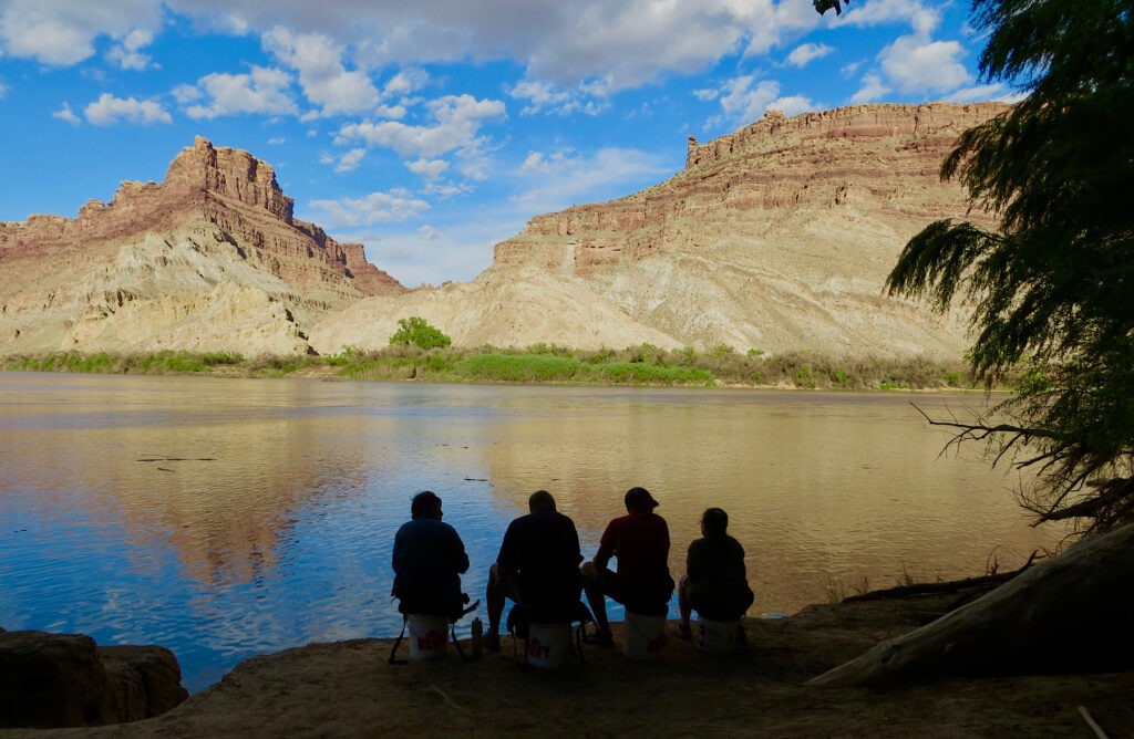 Silhouettes of four people seated above a river with distant cliffs in sunset light.