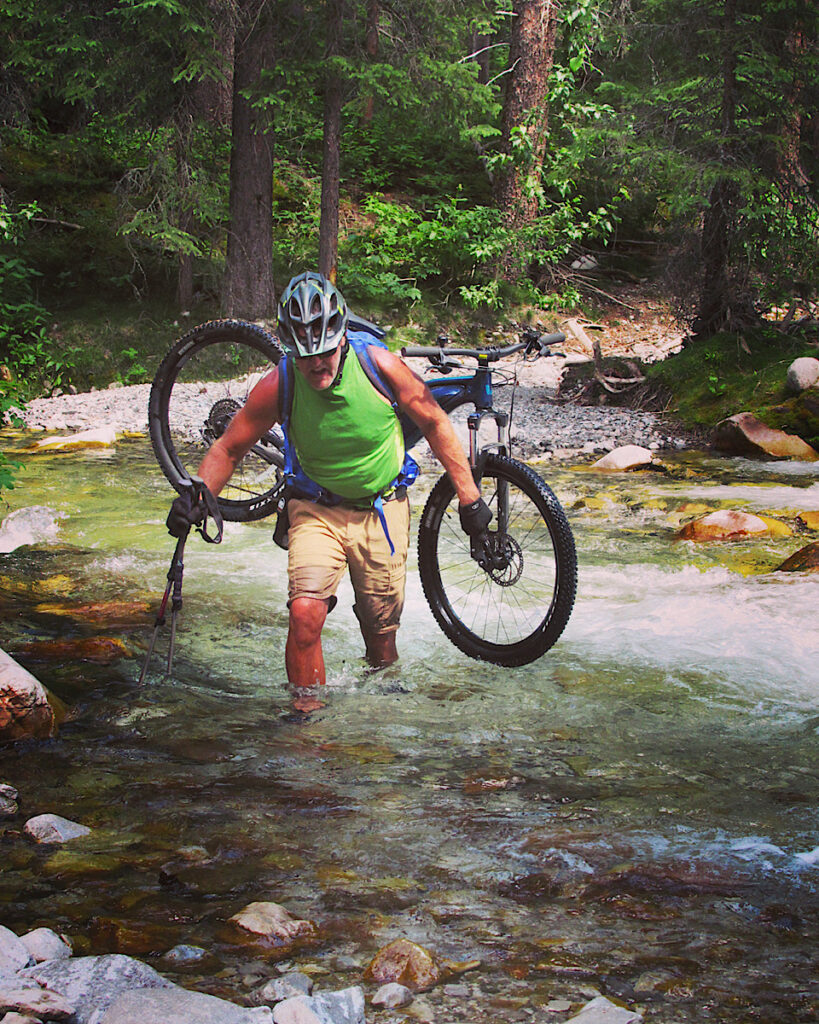 Man in green top and brown shorts carrying bicycle while crossing a large creek.